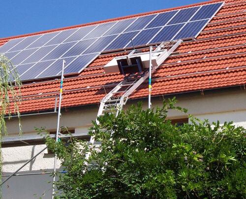 En GEDA Solarlift mot ett tak med en solpanel lastad på flaket. Bredvid liften finns redan monterade solpaneler.